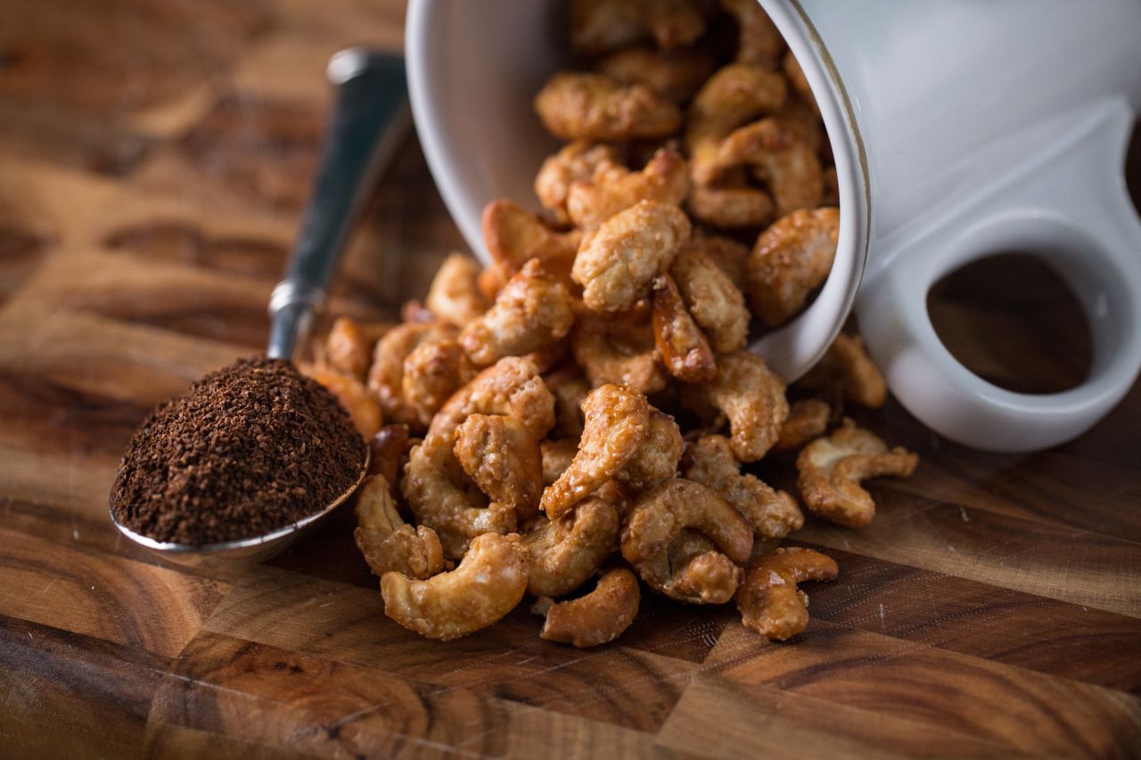 Gourmet coffee stout cashews spilling from a cup next to a spoon of ground coffee on a wooden surface.