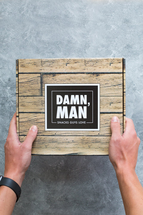 Hands holding a wooden box labeled "Damn, Man: Snacks Guys Love" on a gray background.