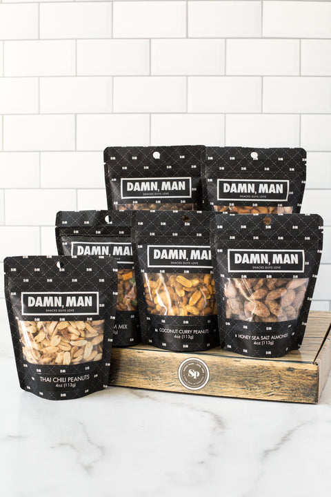 Six bags of "Damn, Man" nuts displayed on a wooden box, featuring a variety of global flavors and classic favorites.