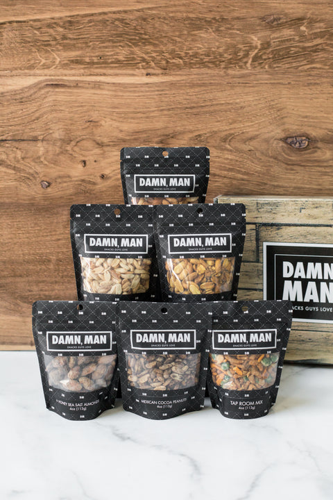 Assorted Damn Man nut bags from the Manly Nut Box on a wooden background, featuring six flavors of hand-crafted, kettle-cooked nuts.