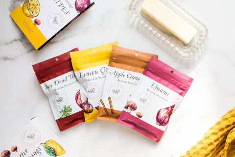 Four-pack of Butter Seasonings with flavors Lemon Garlic, Umami Butter, Sun-Dried Tomato, and Apple Cinnamon next to a stick of butter.