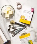 Butter seasonings and recipe book with measuring spoons on marble surface, featuring Lemon Garlic spice pack for cooking and grilling.