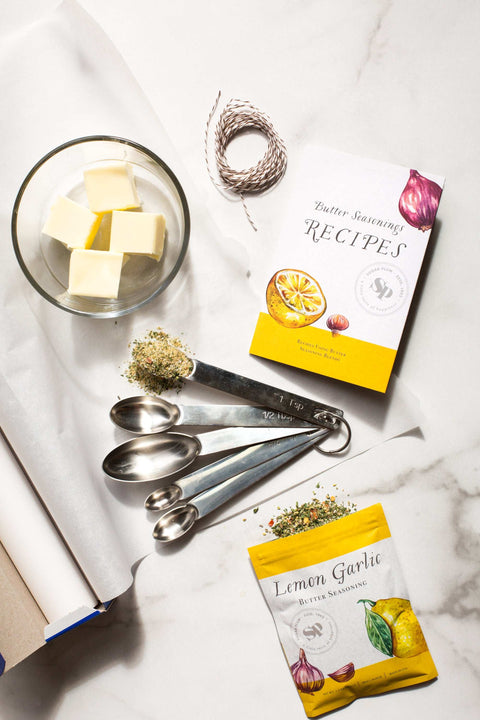Butter seasonings and recipe book with measuring spoons on marble surface, featuring Lemon Garlic spice pack for cooking and grilling.