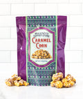 Milk and white chocolate drizzle caramel corn packaging with popcorn scattered around in front of a white tile background.