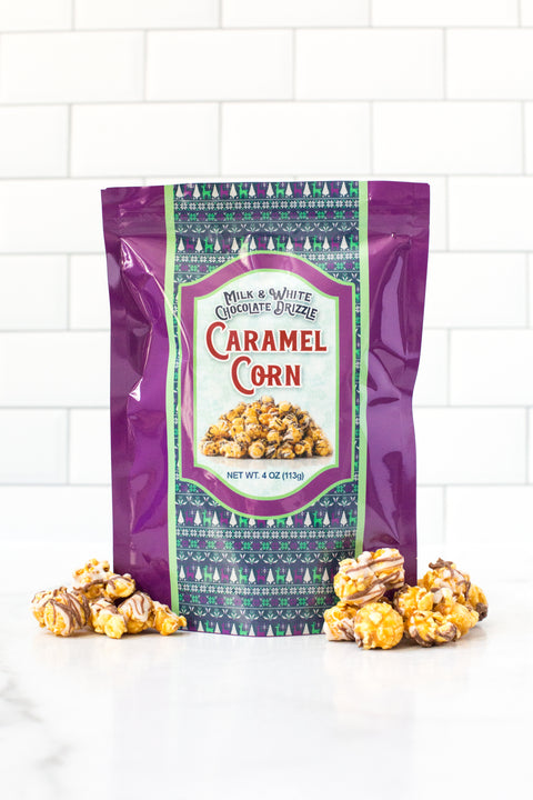 Milk and white chocolate drizzle caramel corn packaging with popcorn scattered around in front of a white tile background.