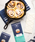 Cinnamon rolls in a skillet with Sugar Plum Cinnamon Roll White Chocolate Bars on a marble table setting.