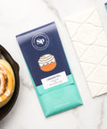 Cinnamon roll white chocolate bars with packaging next to freshly baked cinnamon rolls on a marble surface.