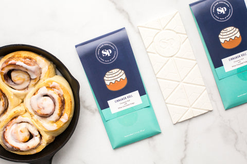 Cinnamon roll white chocolate bars with packaging next to freshly baked cinnamon rolls on a marble surface.