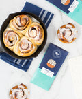 Cinnamon rolls in a skillet with Cinnamon Roll White Chocolate Bars and individual sticky buns on a marble countertop.