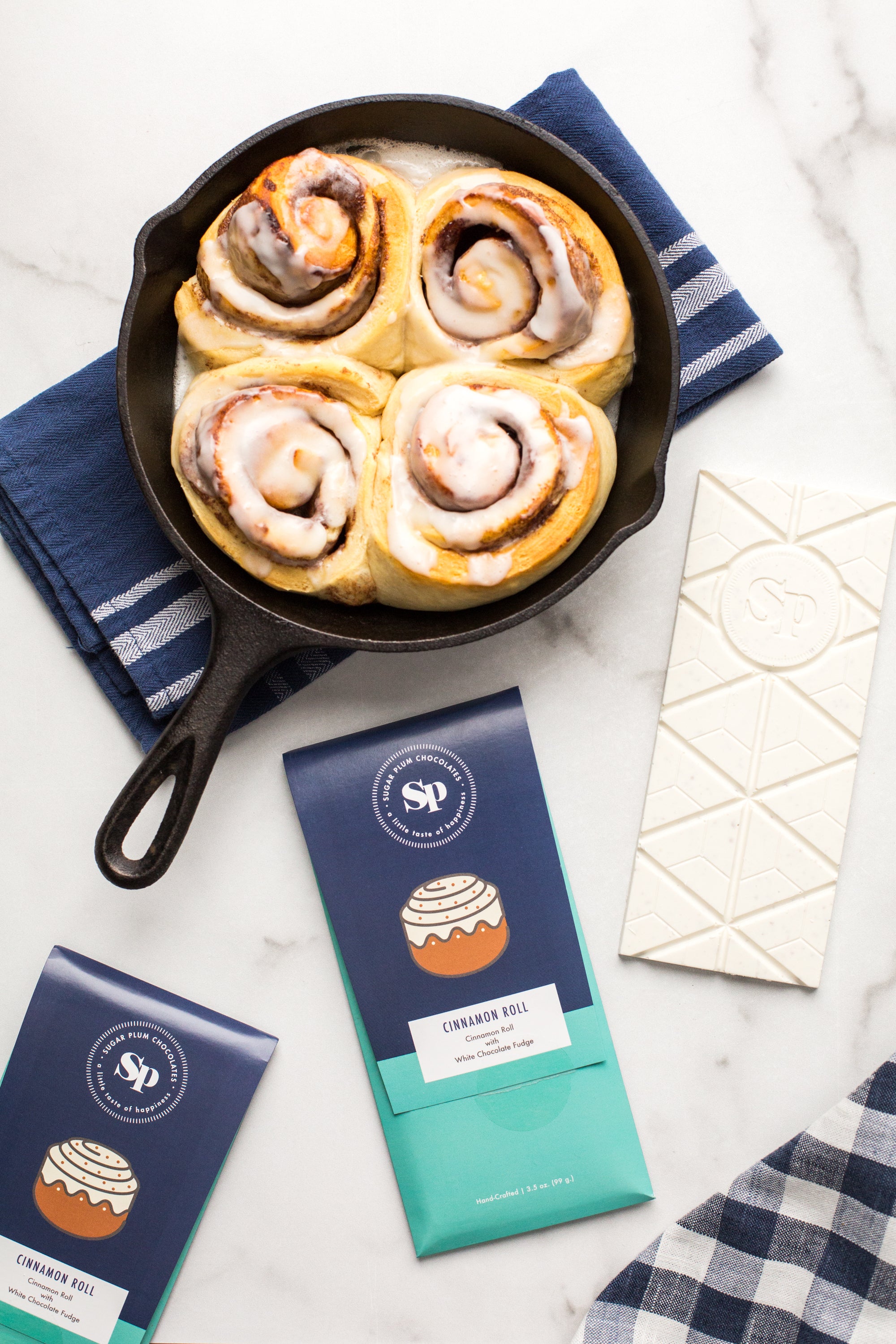 Cinnamon rolls in a skillet with chocolate bars and a checkered napkin on a marble surface.