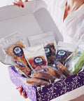Woman holding Sugar Plum Comfort Box filled with 15 gourmet snacks in a purple gift box, perfect for sympathy or get-well gifts.