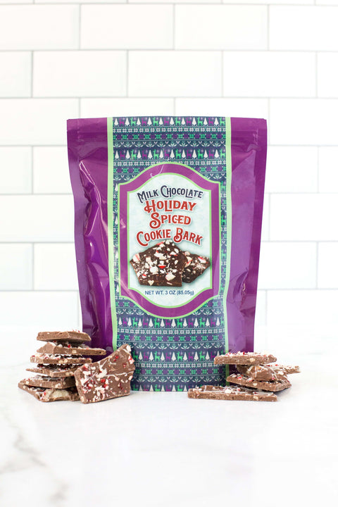 Milk Chocolate Holiday Spiced Cookie Bark with festive packaging and sprinkled chocolate pieces on a white countertop.