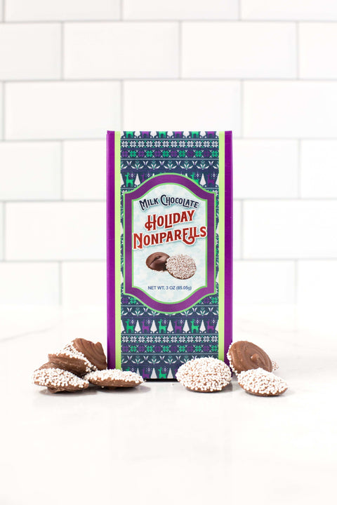 Festive box of milk chocolate holiday nonpareils with scattered peppermint snowflake chocolates on a white tiled background.