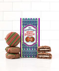 Milk chocolate covered holiday sandwich cookies with festive packaging and stack of cookies on a tiled background