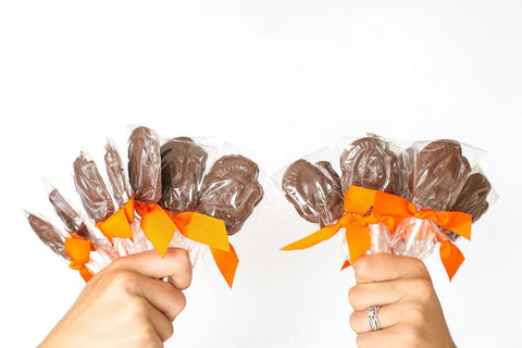Two hands holding 10-pack of handcrafted gourmet milk chocolate turkey lollipops with orange bows, perfect for Thanksgiving.