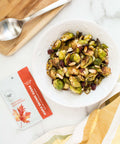 Bowl of roasted Brussels sprouts with nuts, Sugar Plum spice packet, and spoon on wooden board for healthy cooking.
