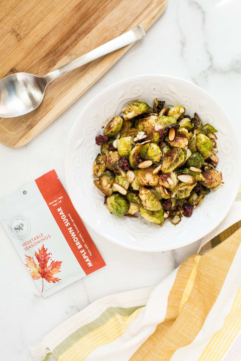 Bowl of roasted Brussels sprouts with nuts, Sugar Plum spice packet, and spoon on wooden board for healthy cooking.