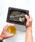 Person holding Beer & Bourbon Liquor Peanuts Tin with a glass of beer, showcasing gourmet roasted peanuts in bold spirit flavors.