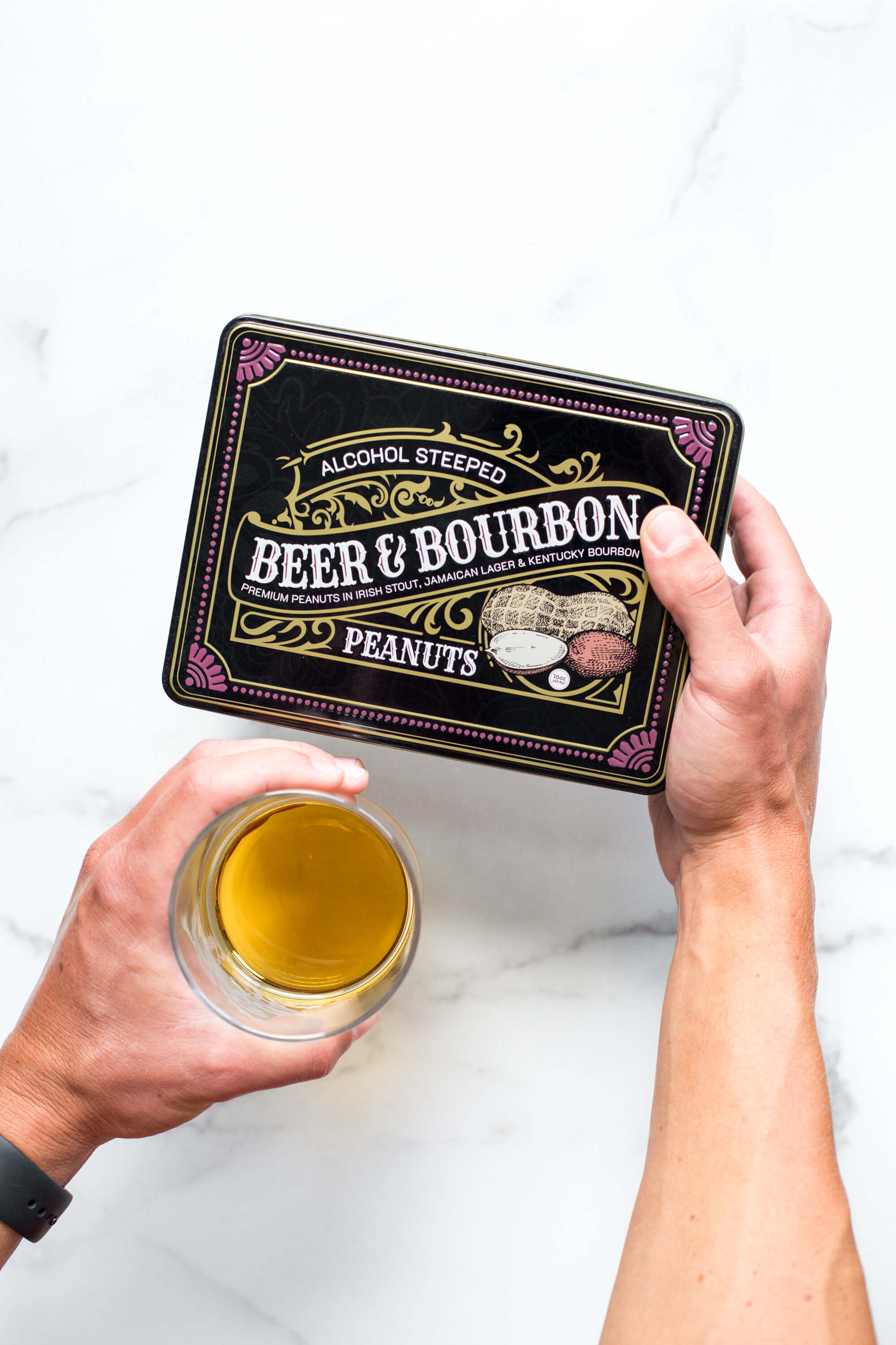 Person holding Beer & Bourbon Liquor Peanuts Tin with a glass of beer, showcasing gourmet roasted peanuts in bold spirit flavors.