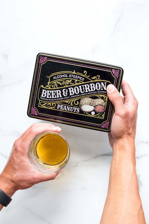 Person holding Beer & Bourbon Liquor Peanuts Tin with a glass of beer, showcasing gourmet roasted peanuts in bold spirit flavors.
