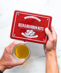 Beer & Bourbon Liquor Nuts Gift Tin with assorted gourmet nuts held above a glass of beer on a marble surface.