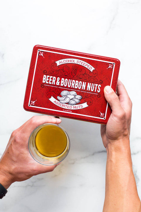 Beer & Bourbon Liquor Nuts Gift Tin with assorted gourmet nuts held above a glass of beer on a marble surface.