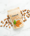 Kettle-cooked pumpkin spice almonds in a beige packaging with illustrated pumpkin, scattered almonds on marble surface.