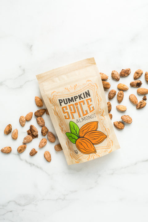 Kettle-cooked pumpkin spice almonds in a beige packaging with illustrated pumpkin, scattered almonds on marble surface.