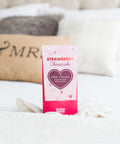 Strawberry Cheesecake White Chocolate Bar in pink packaging on a bed, perfect gourmet dessert gift.