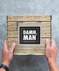 Hands holding a wooden box labeled "Damn, Man: Snacks Guys Love" on a gray background.