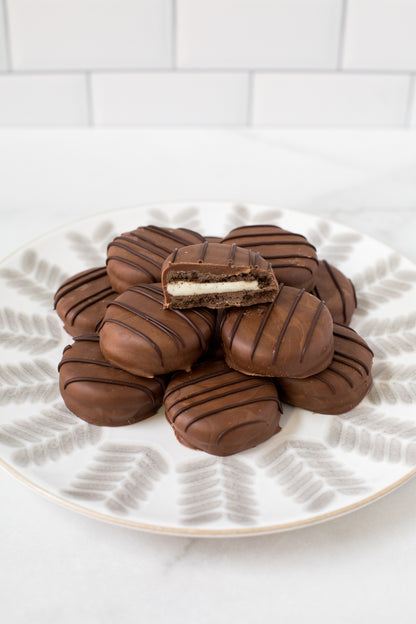 Plate of gourmet chocolate-covered sandwich cookies with a bite taken out of one, showcasing creamy filling and rich chocolate coating.