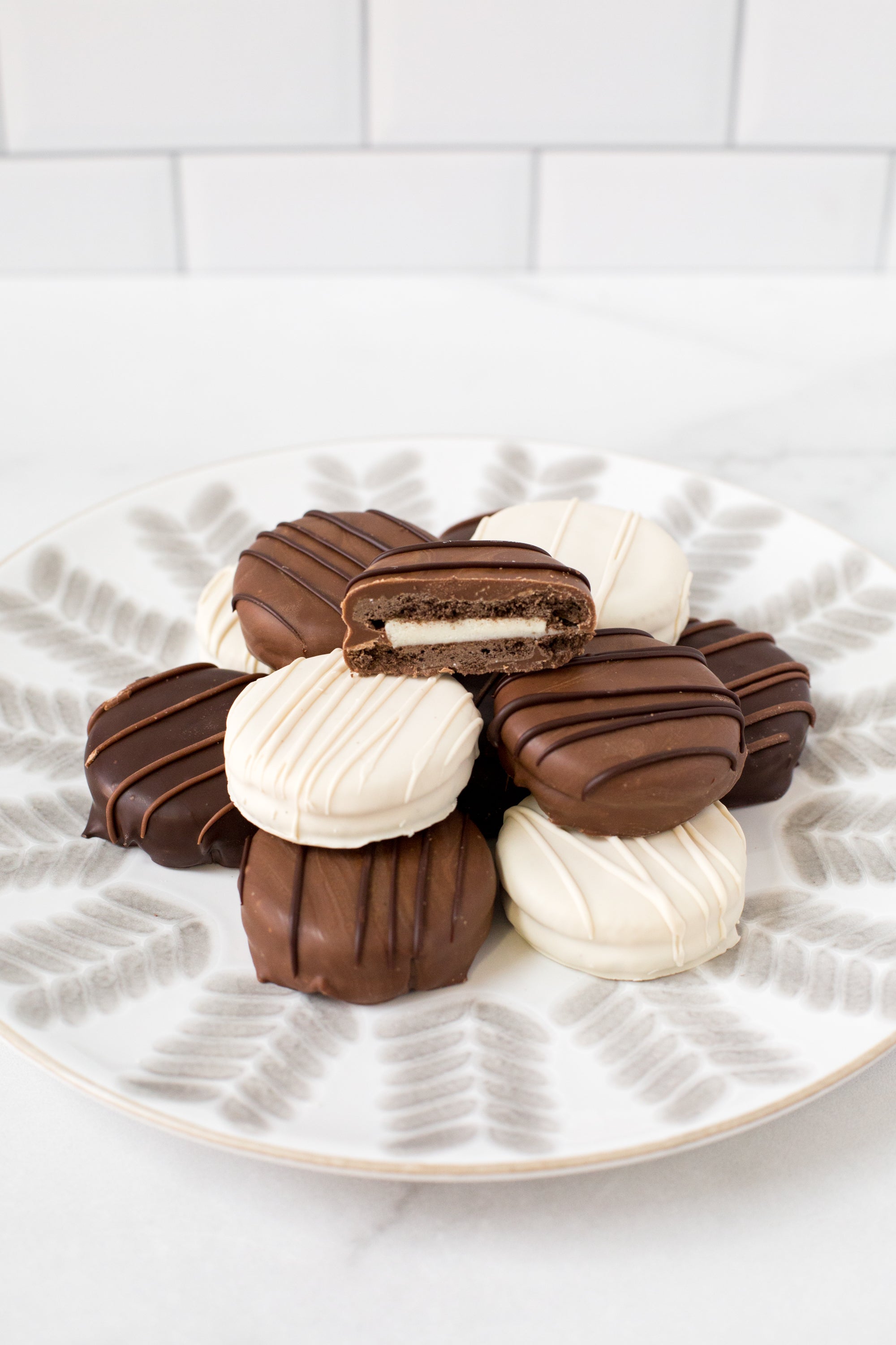 Assorted chocolate-covered sandwich cookies with milk and white chocolate drizzle, arranged on a decorative plate.