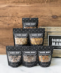 Assorted Damn Man nut bags from the Manly Nut Box on a wooden background, featuring six flavors of hand-crafted, kettle-cooked nuts.
