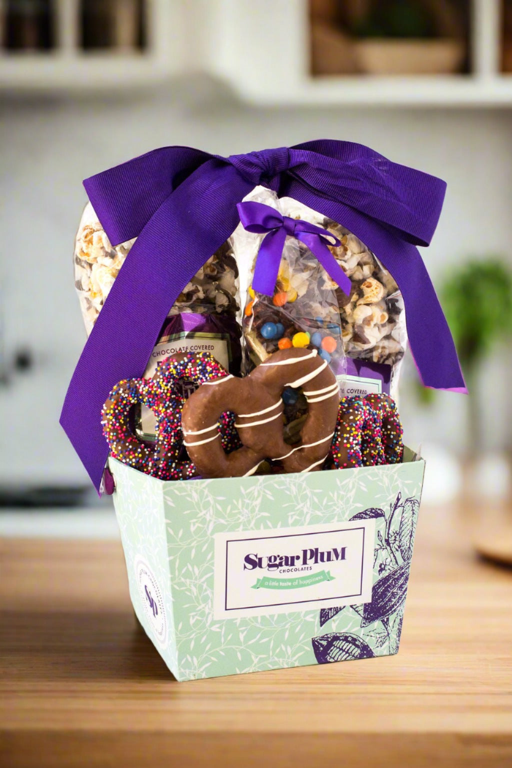 Gourmet chocolate gift basket with purple ribbon featuring cookies, popcorn, and pretzels by Sugar Plum. Ideal for special occasions.