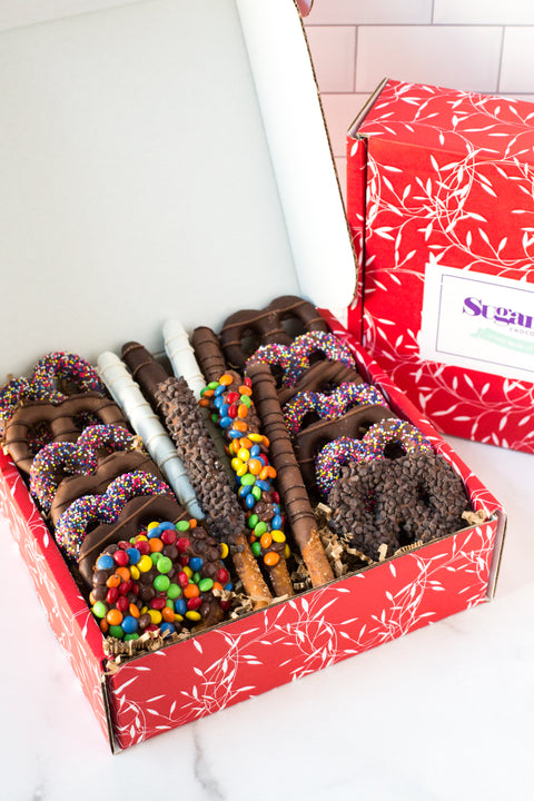 Open box of Sugar Plum's Chocolate Pretzel Passion Gift Tray that includes 22 pieces of assorted chocolate-dipped pretzels, including pretzel logs covered in chocolate candy pieces, chocolate sandwich cookie logs, double chocolate chip pretzels, nonpareil pretzels, candied chocolate gem pretzels, and pretzels dipped in signature milk, dark, and white chocolates.
